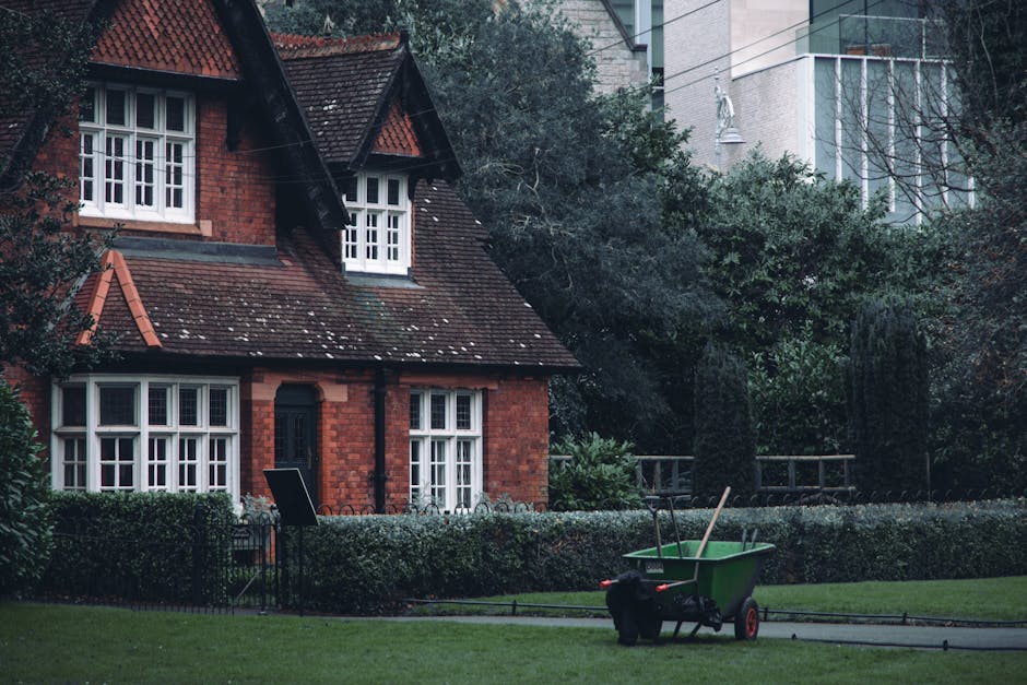 A traditional red-brick house with a pitched, tiled roof and white-framed casement windows, surrounded by a well-maintained garden. In the foreground, a green wheelbarrow filled with gardening tools s